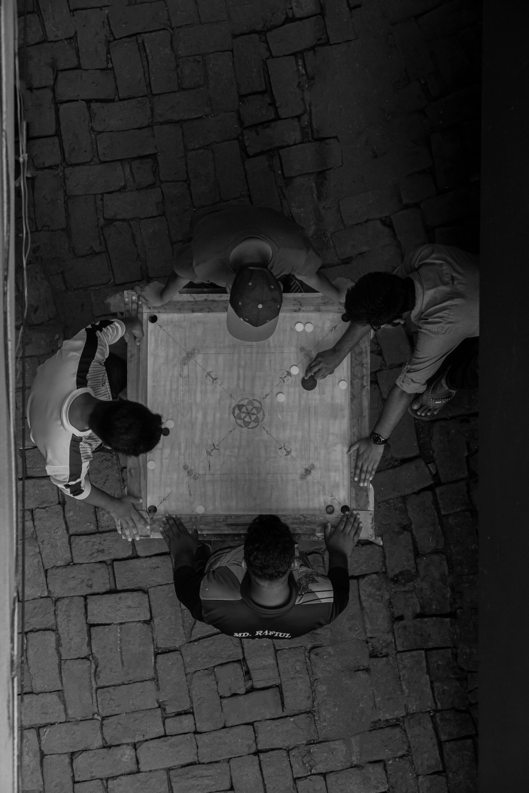 Top view of four men playing carrom on a brick pavement, captured in black and white.